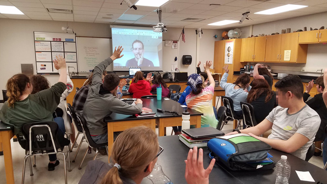 High school students in a classroom look towards a projector wall that shows Toni on a video call, some students are raising their hands in response to a question